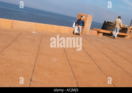 Eine Frau sitzen trinken Mate Kräutertee und ein Mann zu Fuß auf dem Bürgersteig, auf dem Spaziergang am Flussufer am Meer entlang des Flusses Rio De La Plata Ramblas Sur, Gran Bretagna und Republica Argentina Montevideo, Uruguay, Südamerika Stockfoto