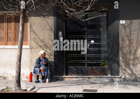 Eine alte Frau sitzt auf dem Bürgersteig trinken Mate Kräutertee, mit einer roten Warmwasser Thermoskanne vor einem schwarzen Eisentor Tor. Montevideo, Uruguay, Südamerika Stockfoto