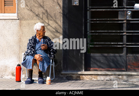 Eine alte Frau sitzt auf dem Bürgersteig trinken Mate Kräutertee, mit einer roten Warmwasser Thermoskanne vor einem schwarzen Eisentor Tor. Montevideo, Uruguay, Südamerika Stockfoto