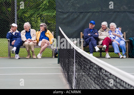 Ältere Menschen im Ruhestand, Männer und Frauen sitzen auf Bänken neben einem Tennisplatz net gerade ein Spiel im Gange Stockfoto