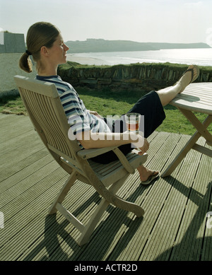 Eine Frau sitzt auf einem Liegestuhl an Deck in der Sonne mit Blick auf Fistral Strand lächelnd Stockfoto