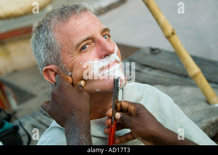 Horizontale Nahaufnahme eines westlichen Touristen, die eine Rasur von einem lokalen indischen Friseur außerhalb an den Ufern des Flusses Ganges. Stockfoto