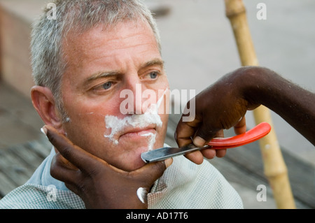 Horizontale Nahaufnahme eines westlichen Touristen, die eine Rasur von einem lokalen indischen Friseur außerhalb an den Ufern des Flusses Ganges. Stockfoto