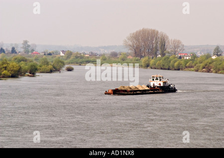 Ein Transportschiff mit Sand auf der Weichsel in Polen, umgeben von üppigen grünen Ufern und weit entfernten Häusern unter einem trüben Himmel. Stockfoto