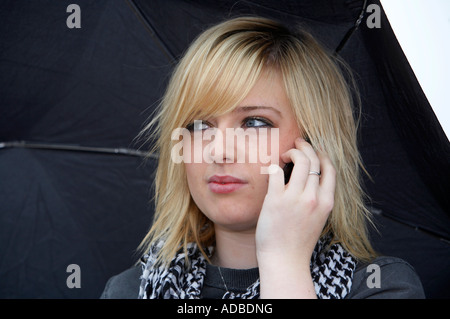 18 jährige Blondine hält Regenschirm suchen wütend Gespräch auf dem Handy hören Stockfoto