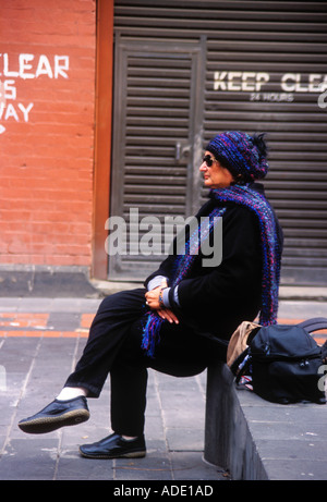 Ältere Frau im Winter Kleidung und Sonnenbrille sitzt auf konkrete Bank mit Ziegel Wand Spruch klar Melbourne Victoria Aust halten Stockfoto