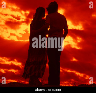 Voller Länge Blick auf junges Paar Arm in Arm Silhouette gegen brillanten Wolkenformationen bei Sonnenuntergang Stockfoto