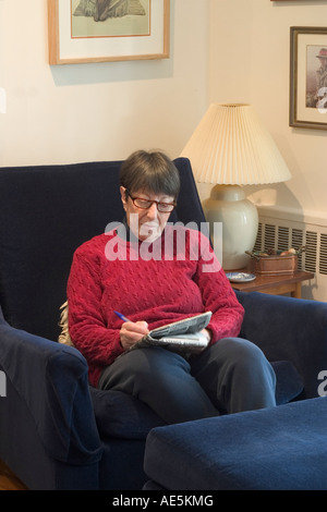 Ältere Frau 70 sitzen in einem gemütlichen Wohnzimmer Stuhl machen die New York Zeiten Kreuzworträtsel in Stift Stockfoto
