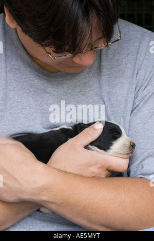 Chinesisch-amerikanischen Mann zärtlich wiegt einen zwei Wochen alten Mischling Welpe schlafend in seinen Armen Stockfoto