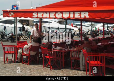 Kellner und Kunden in der beliebten Senequier Patisserie und der roten Bar auf dem Bürgersteig am Wasser mit Motorbootstarts hinter St. Tropez in Südfrankreich Stockfoto