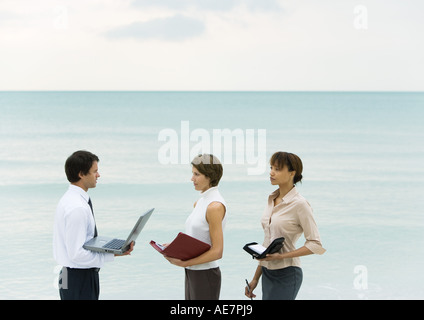 Geschäftsmann mit Laptop, Frauen halten Tagesordnungen, Meer im Hintergrund Stockfoto