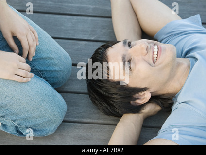 Junger Mann mit Händen hinter dem Kopf, auf dem Boden liegend lächelnd, jemand kniete neben ihm, Teilansicht Stockfoto