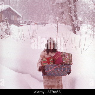 Weihnachtsszene mit attraktiven jungen Frau gekleidet herzlich zu Fuß Weg durch Schneeverwehungen tragen Armload Geschenke Stockfoto