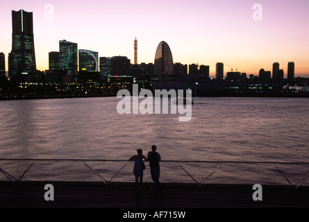 Ein Paar am Osanbashi Pier bei Sonnenuntergang mit Yokohama Skyline bei Sonnenuntergang, zeigt Landmark Tower (links) Cosmo Clock (Mitte) und Grand Intercontinental Hotel (rechts) Kanagawa, Japan Stockfoto