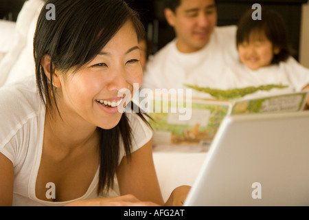Mutter arbeitet auf Laptop-Computer im Bett während Vater auf den Sohn im Hintergrund liest Stockfoto