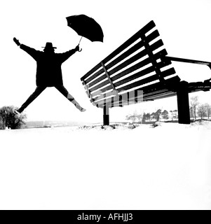 Eine Person mit einem Regenschirm springt nach einem Schneefall neben einer Parkbank in die Luft. East Sussex, Großbritannien. Stockfoto