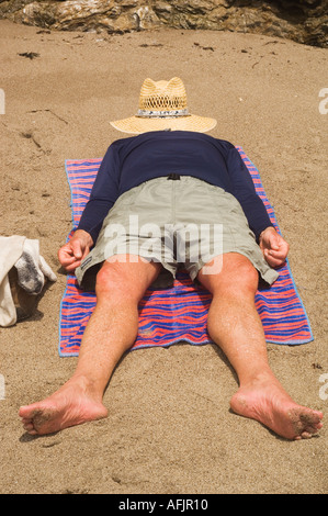Mann liegend auf einem Handtuch am Strand mit einem Strohhut bedeckte sein Gesicht barfuß in kurzen Hosen und ein langärmeliges Hemd Stockfoto
