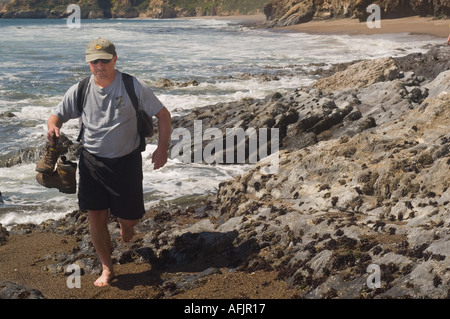 Mann in shorts ein t Shirt und Basecap tragen Wandern Stiefel und tragen einen Rucksack zu Fuß über Barnacle Felsen bedeckt Stockfoto