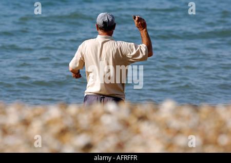 ein Mann stand auf einem Kieselstrand mit seinen Arm hob werfende Steinen ins Meer mit dem Wasser im Hintergrund an einem Sommertag Stockfoto