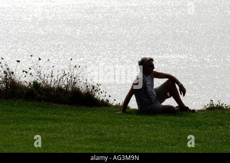 junger Mann sitzt auf Klippen bei Sonnenuntergang mit Blick auf das glitzernde Meer den Sonnenuntergang Stockfoto