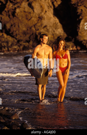 Westseite des Paares auf dem Strandspaziergang am Rand des Wassers bei Sonnenuntergang auf Maui. Der Mann trägt ein Surfbrett. Stockfoto