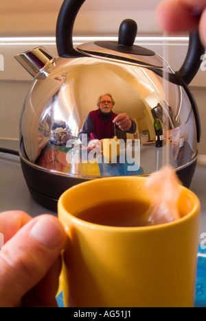 Mann, der eine Tasse Tee macht, reflektiert in einem glänzenden Edelstahl-Wasserkocher Stockfoto