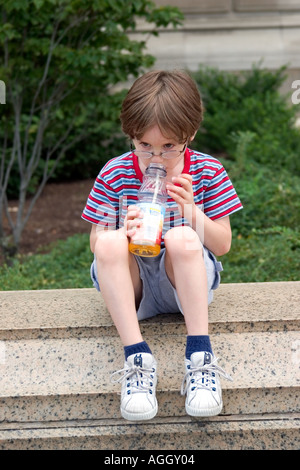 kleiner Junge im gestreiften Hemd sitzt auf konkrete Stützmauer Zaun trinken eine Flasche Apfelsaft durch einen Strohhalm Stockfoto