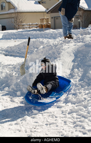 Kleiner Junge Rodeln bergab im Schnee während seiner Eltern im Hintergrund schaut Stockfoto