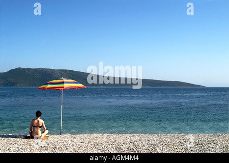 Ein Sonnenanbeter sitzen unter einem Sonnenschirm an einem Kiesstrand, auf der Insel Alonissos in Griechenland. Stockfoto