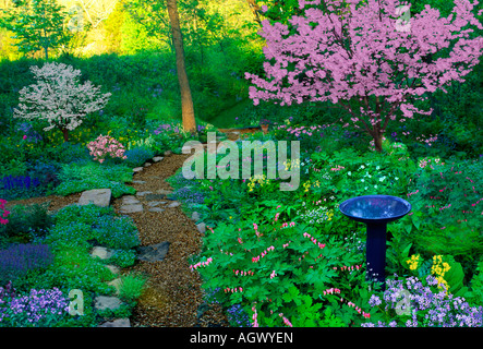 Aggregierte Bahnkurven im Frühjahr schattieren, Garten mit Vogeltränke und vielen blühenden Blumen und rosa und weiße Hartriegel Bäume Stockfoto