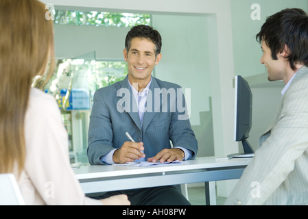 Junges Paar sitzt auf Schreibtisch von lächelnden Geschäftsmann, Unternehmer im Fokus Stockfoto