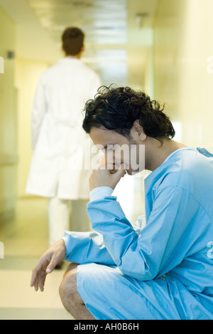 Männliche Patienten im Krankenhausflur hockend, Kopf, blickte, Arzt zu Fuß entfernt, im Hintergrund Stockfoto