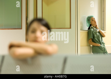 Medizinische Arbeiter stehen im Wartezimmer des Krankenhauses, die Hände in den Taschen, Blick in die Kamera, Frau ruht Kopf auf Arme im Vordergrund Stockfoto