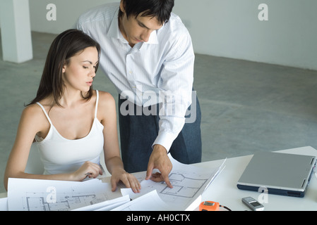 Frau sitzt am Tisch, Blick auf Blaupausen, Mann stehend, über die Schulter schauen Stockfoto