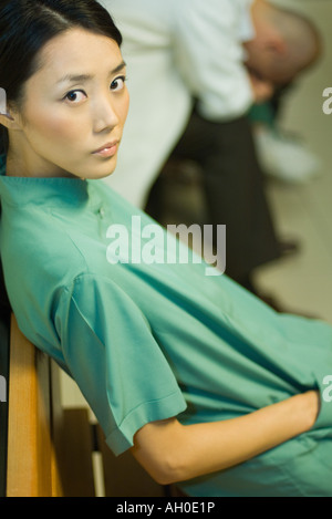 Medizinische Arbeitnehmerin sitzend mit Hand in der Tasche, Blick in die Kamera, Arzt mit Kopf nach unten im Hintergrund Stockfoto