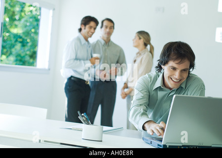 Geschäftsmann im Büro, mit Laptop-Computer, lächelnd, Mitarbeiter im Hintergrund Stockfoto
