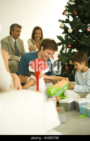 Mann und zwei Kindern öffnen Weihnachten präsentiert vor Weihnachtsbaum, Familie, die gerade im Hintergrund Stockfoto