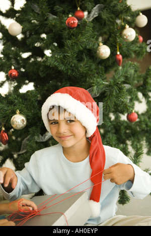 Junge Weihnachtsgeschenk vor Weihnachtsbaum, Santa Hut trägt, lächelnd in die Kamera zu öffnen Stockfoto