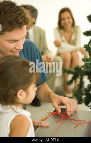Mann und Mädchen Weihnachtsgeschenk, Erwachsene im Hintergrund öffnen Stockfoto