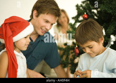 Vater und zwei Kinder öffnen Weihnachten präsentieren vor Weihnachtsbaum, Mädchen trägt Weihnachtsmütze Stockfoto