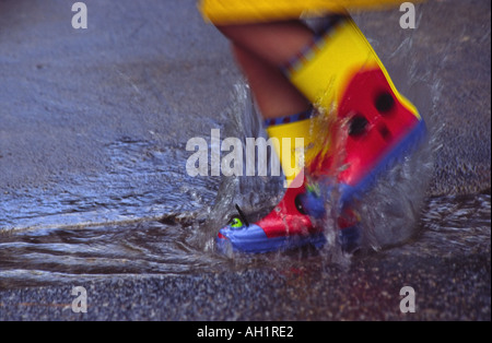 Nahaufnahme eines Kindes in bunten Regenstiefel oder Gummistiefel in einer Pfütze auf der Straße durch Spritzwasser Stockfoto