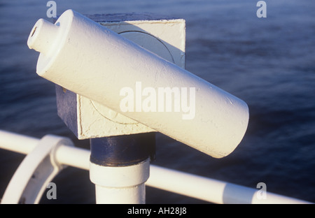 Nahaufnahme eines Teleskops am Meer neben weißen Geländer mit Blick auf eine Schwellung Meer in warmes Sonnenlicht Stockfoto