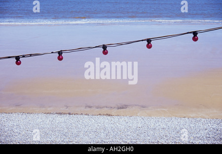 Eine Reihe von roten Glühbirnen aufgereiht über einen nassen Sandstrand mit einem Streifen aus grauen Kieseln und ruhigen blau graue Meer mit Wellen plätschern Stockfoto