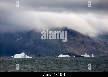 Dramatische Berge unter schweren niedrigen grauen Himmel mit weißen Eisberge schwimmt im Gegensatz dazu auf See vor der südwestlichen Küste Grönlands Stockfoto