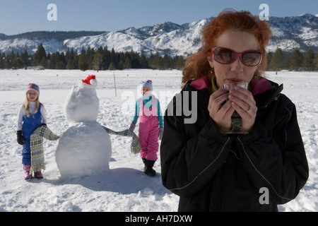 Reife Frau mit einem Glas mit ihren beiden Töchtern stehen neben einem Schneemann Stockfoto