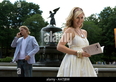 Junge Frau hält ein Buch mit einem jungen Mann telefonieren mit einem Handy hinter ihr, Central Park, Manhattanrk Stadt Stockfoto