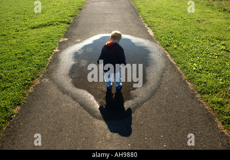 kleiner Junge blickte auf Pfütze und Schatten entlang einem Gehweg Stockfoto