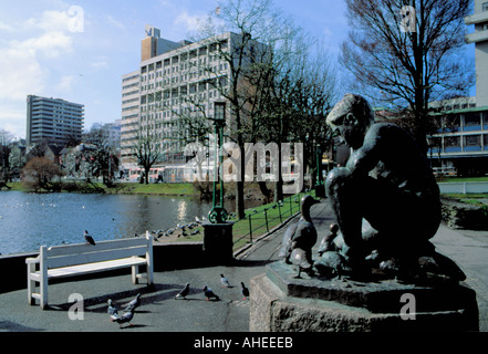 Bronzeskulptur von jungen füttern Enten, mit dem Atlantic Hotel darüber hinaus über Breiavatnet, Stavanger, Rogaland, Norwegen gesehen. Stockfoto