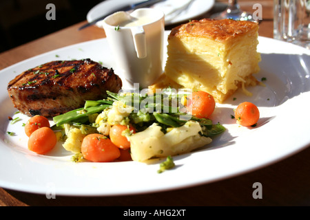 Gegrilltes Schweinefleisch, serviert mit cremigem Kartoffelgratin, gedämpftem Gemüse und einer Seite cremiger Soße auf einem weißen Teller. Stockfoto