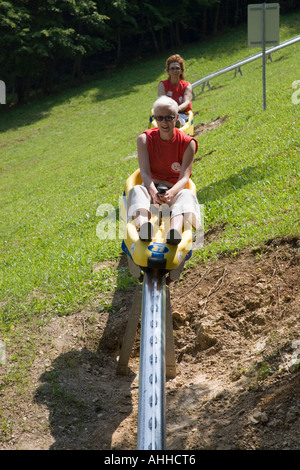 Damen saßen Reiten auf gelben Schlitten herab, Sommerrodelbahn fahren auf Metallschiene unteren Speisekarte Skipiste in Bled Slowenien Stockfoto